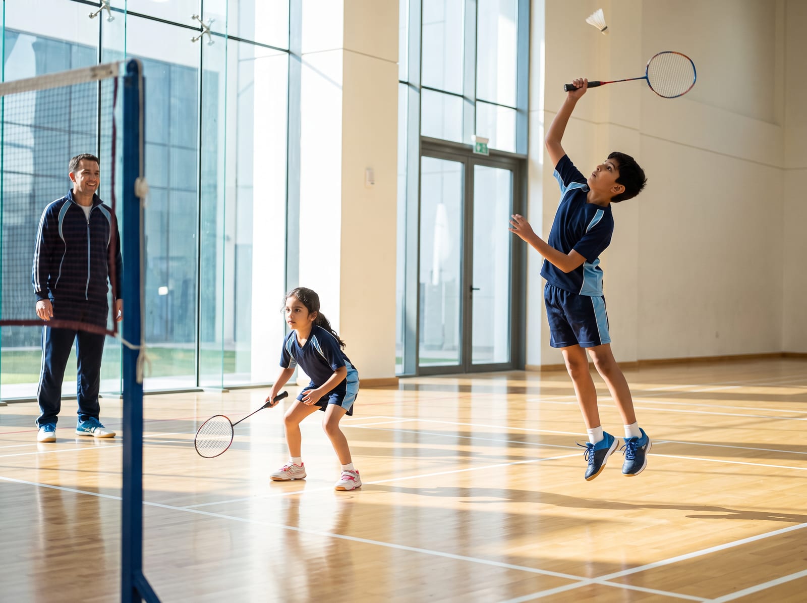 A child leaping to smash a shuttlecock in a bright indoor badminton hall while a teammate and a coach look on